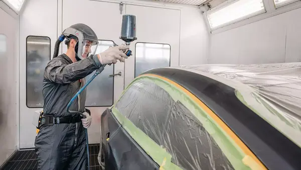 Autolackierer trägt in Lackierkabine Schutzanzug und lackiert ein vorbereitetes Auto.