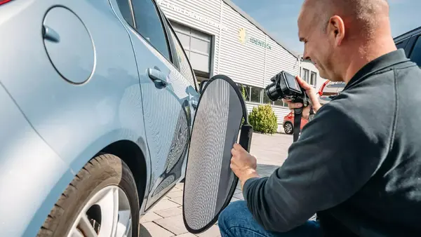 Mann prüft mit Kamera und Lackschaden-Reflektor ein Auto auf Dellen und Karosserieschäden.