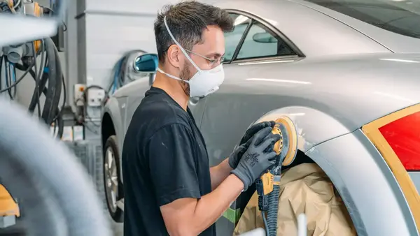 Automechaniker schleift mit Schutzmaske und Handschuhen Kotflügel eines Autos in Werkstatt.